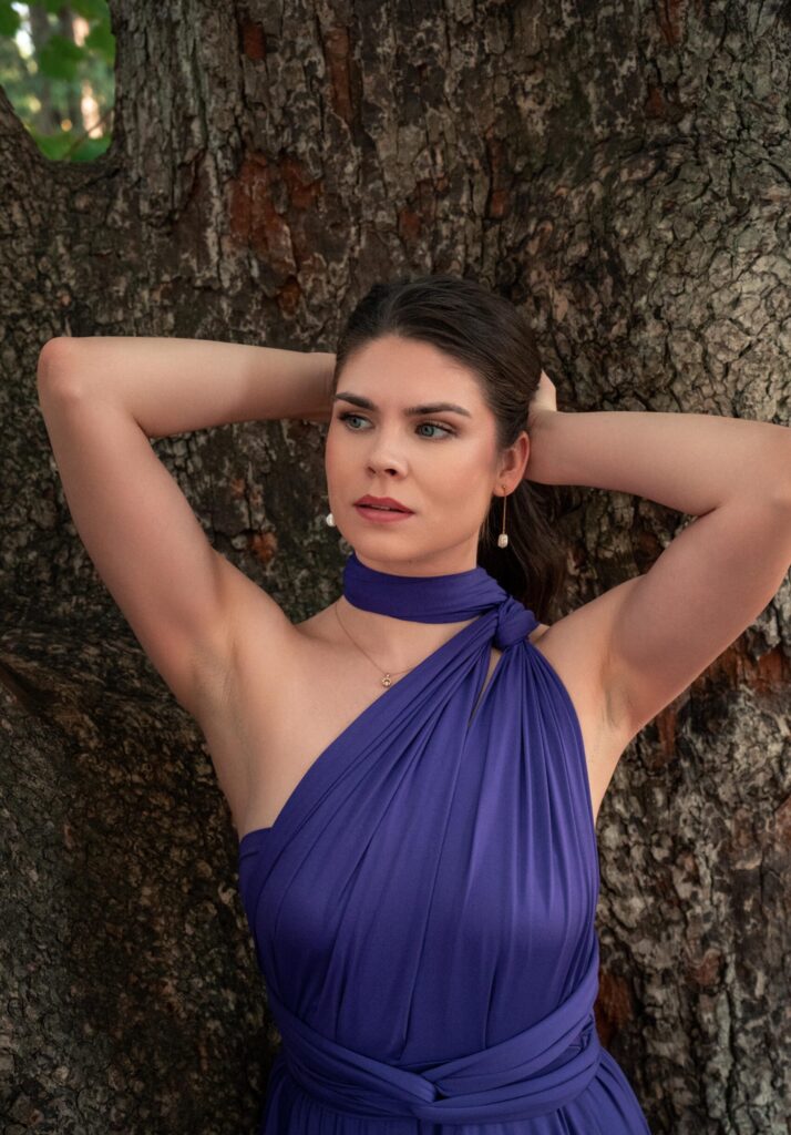 Frau im violetten One-Shoulder-Wickelkleid vor Baumstamm, Hände im Nacken, Portrait