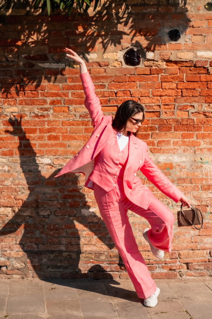 Frau im pinken Hosenanzug in dynamischer Pose vor Ziegelmauer, Handtasche und weiße Sneaker