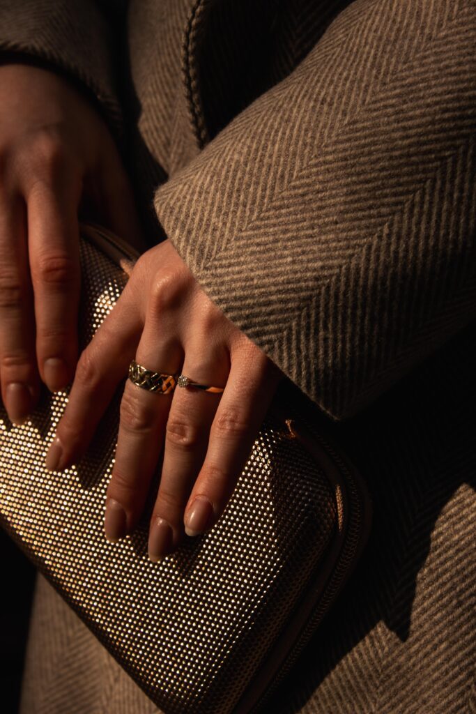 Detail: Hände mit Goldringen auf goldener Clutch, Ärmel eines Fischgrät-Mantels im Sonnenlicht