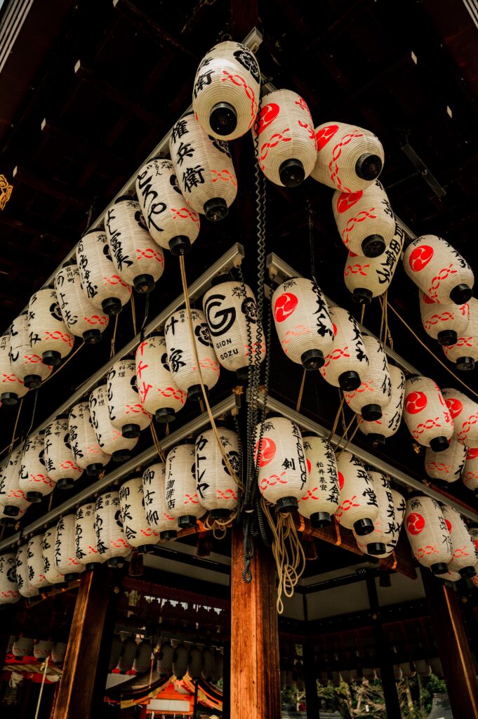 Viele weiße Papierlaternen mit japanischer Schrift und roten Symbolen unter Holzdach