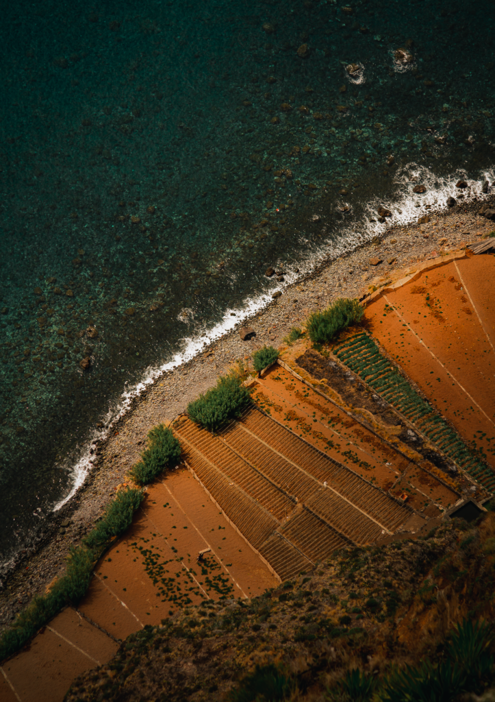 Luftaufnahme: türkises Meer, Kiesstrand und terrassierte rötliche Felder an der Küste, Madeira