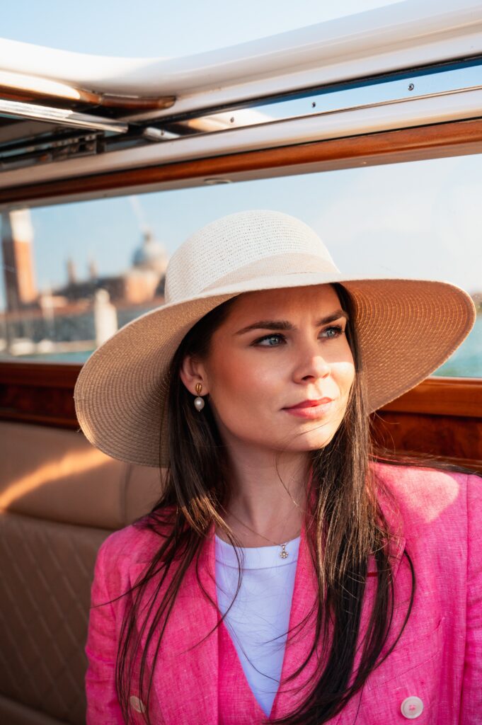 Frau mit breitem Strohhut und pinkem Blazer in einem Boot, Venedig im Hintergrund